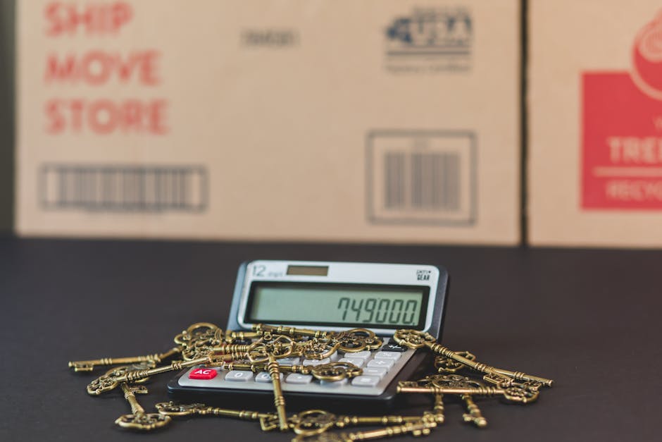 A close-up image shows a small calculator displaying the number 749800 on its screen, situated on a black surface. Scattered across the calculator are numerous brass keys, varying in size and style, some with intricate designs, indicating they may be vintage or decorative keys. Behind the calculator and keys, several large cardboard moving boxes are visible, with printed labels including barcodes, handling instructions like 'Ship Move Store,' and other shipping details. The boxes are positioned in a row, with some overlapping, and are likely part of a home relocation process facilitated by Removals Harrow, a professional removals service. The environment appears to be indoors, with soft lighting highlighting the objects, emphasizing a scene associated with packing, moving, and furniture transportation as part of house removal activities.
