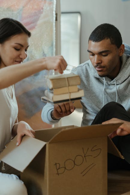 Two individuals, a woman and a man, are engaged in packing books for a home relocation. The woman is holding a small stack of books, while the man is passing her a single book wrapped with elastic ban