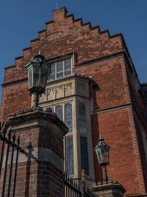 Photograph showing the exterior of a historic red brick building with a steeply stepped gable and a stone bay window featuring decorative leaded glass panes. At the front, there are two black metal la