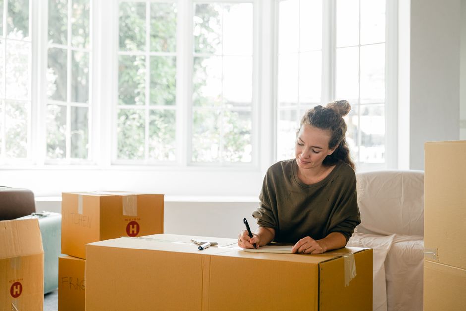 A woman signing a packing list or inventory form while seated at a table inside a well-lit room with large windows and white walls. Surrounding her are several cardboard moving boxes, some sealed with packing tape and labeled, indicating the process of home relocation. The boxes vary in size and are made of brown corrugated cardboard, with a few showing visible packing materials like plastic wrap or fabric surrounding items inside. She is wearing a casual, dark-colored long-sleeve top and has her hair tied back in a bun. The setting suggests an organized packing and furniture transport operation, and this scene exemplifies the logistics involved in a professional house move, with Removals Harrow providing safe and efficient packing and moving services as part of this relocation process.