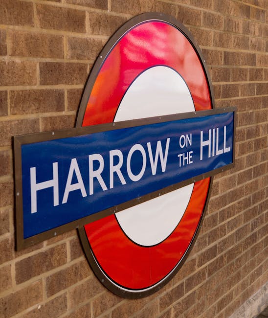 A close-up image of a circular London Underground sign mounted on a brick wall, featuring a red outer ring with a white central circle and a blue rectangular bar across the center displaying the text 'HARROW ON THE HILL' in white capital letters. The sign is part of a house removal and home relocation process, with visible hardware securing it to the wall. The surrounding environment appears to be an indoor or semi-indoor setting, possibly at a station or property entrance, with controlled lighting illuminating the sign and the brick wall behind it. This image supports context related to transportation and moving logistics, common aspects of furniture transport and packing during house removals, as handled by professionals like Removals Harrow.