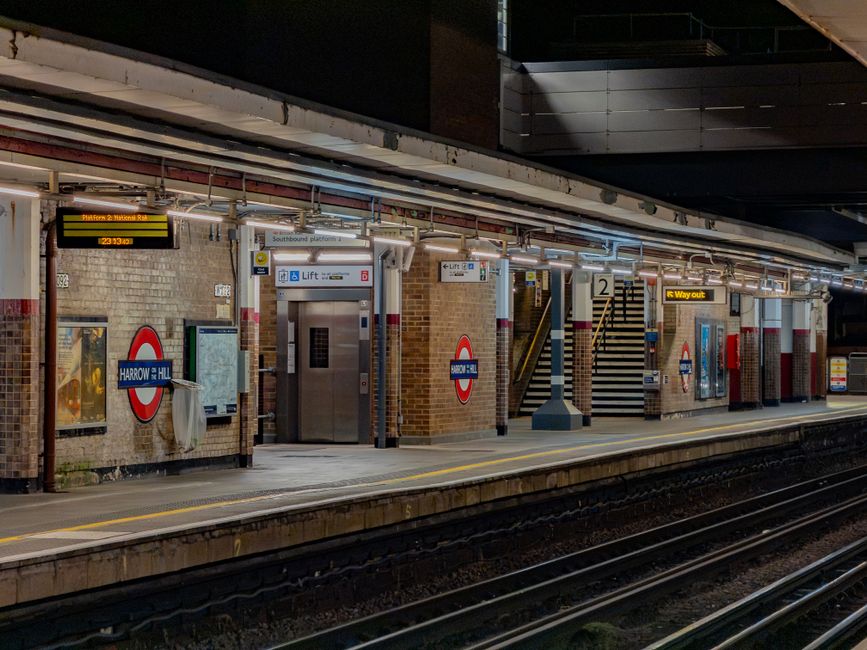 A young man sits on a black metal bench on the platform of Harrow on the Hill underground station, part of the London Underground network, located in Harrow. Behind him, a white and red Tube train wit