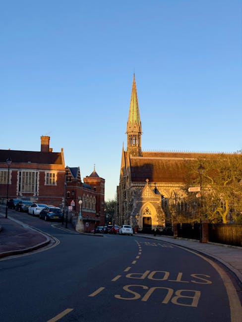 A daytime street scene featuring a narrow road with a 'BUS STOP' marking and a yellow central line, situated in a historic area of Harrow on the Hill. On the left, a row of parked cars lines the pavement alongside red-brick buildings, some with pitched roofs and chimneys. On the right, a large, historic stone church with a tall, pointed spire dominates the scene; its Gothic architectural details include arched windows and a decorative doorway. The sky above is clear and blue, providing natural lighting that highlights the textures of the buildings and street. The location suggests a typical urban environment where house removals and local logistics might involve careful navigation of narrow streets and consideration of historic landmarks, relevant to services offered by Removals Harrow in the context of house relocation or furniture transport in areas with limited street width.