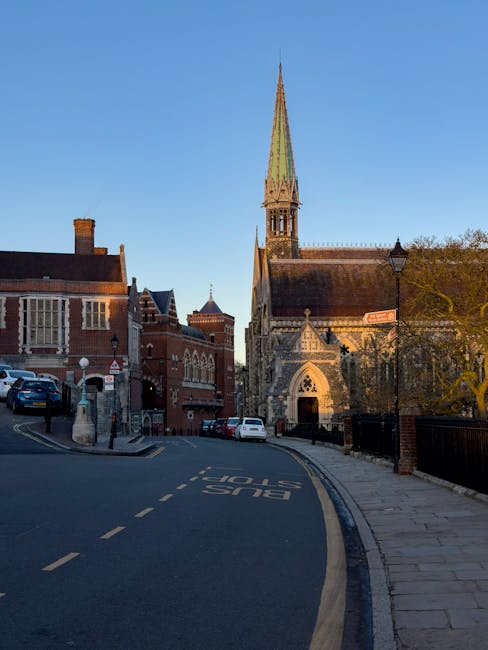 A quiet residential street in Harrow with traditional brick houses featuring pitched roofs, white-framed sash windows, and brick chimneys. The street is lined with pavement, a black metal lamppost, and small front gardens with fencing and greenery. A silver car is visible driving along the narrow, curved road, which is suitable for home relocation and furniture transport services. On the right, a brick house with white decorative columns and steps leading to the entrance is partially visible, indicating an area where professional removal companies such as Removals Harrow might assist with packing and moving. The environment appears clean and peaceful, with a large tree providing greenery, and the overall scene reflects the typical setting for house removals on narrow streets, emphasizing careful loading and transportation processes in an urban residential context.