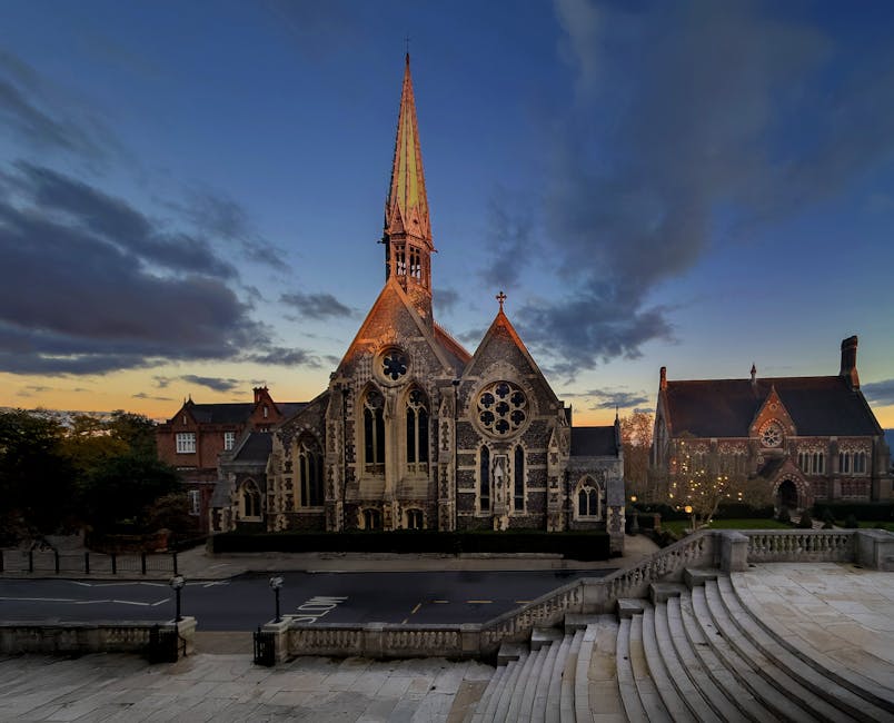 A large Gothic-style stone church with a tall, pointed spire, located on a wide paved area with curved steps leading up to an entrance. The church features intricate stained glass windows, including circular and pointed-arch designs, and decorative stonework. The surrounding environment includes indoor and outdoor space with street lamps illuminating the area, decorative fencing, and nearby trees. The sky above is partly cloudy during the early evening or late afternoon, casting a warm light on the building. This scene appears to be part of a churchyard or historic site in Harrow, and the image is associated with a house removals and packing guide for Harrow, highlighting the importance of careful packing and transportation of furniture and personal belongings during a home relocation, as coordinated by Removals Harrow, a professional removals service specializing in comprehensive moving logistics.