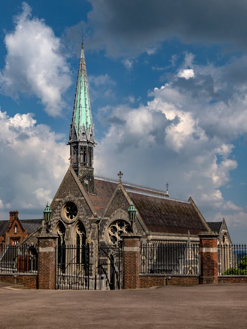 A historic stone church with Gothic-style architecture, featuring a tall, pointed spire with a greenish patina, large arched stained glass windows, and decorative stone carvings. The church is enclosed by a black wrought iron fence with brick pillars at the entrance, set on a paved area. The sky above is partly cloudy with patches of blue, and the surrounding scene suggests an outdoor setting in daylight, illustrating the type of building involved in house removals or packing projects supported by Removals Harrow as part of residential relocation services.