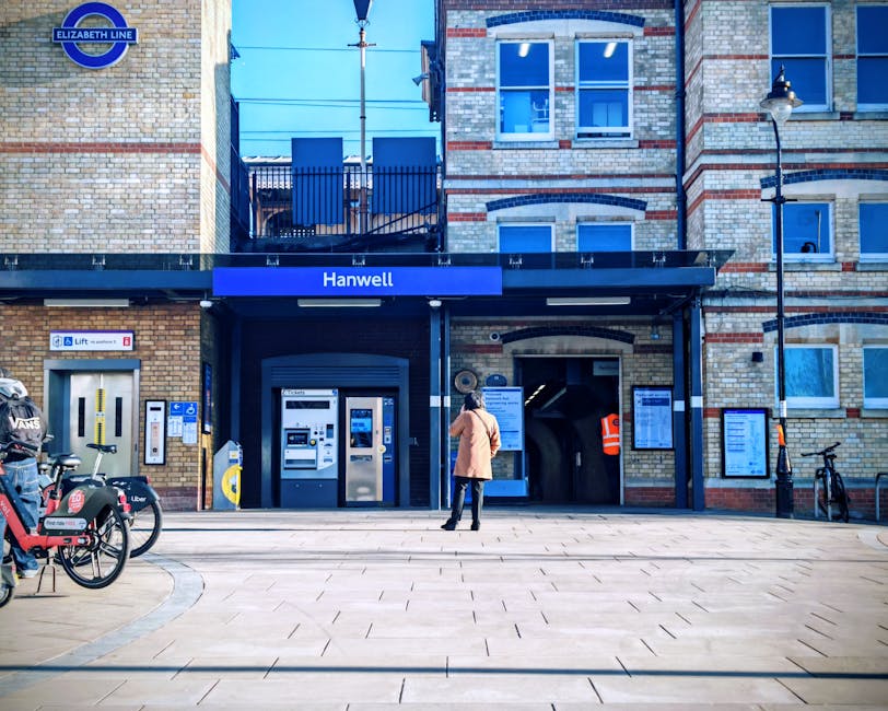 The image depicts the exterior entrance of Hanwell station, part of the Elizabeth Line, with a person standing on the pavement facing the station entrance. The station features a brick facade with large windows and a prominent blue sign displaying the station name. In front of the station, there are several bicycles parked on bike racks, one with a Red VANS bag attached. To the left, a small section of ticket machines and an automated entry gate are visible, with clear signage indicating access points and ticketing options. The pavement is spacious with light-colored tiles, and a streetlamp is positioned on the right side of the image. Above the station entrance, there is an overhead canopy protected by a metal railing, providing shelter for passengers. The scene is well-lit with natural daylight, and the overall setting illustrates an urban transport hub suitable for home relocation or furniture transport during moving services, as offered by Removals Harrow.