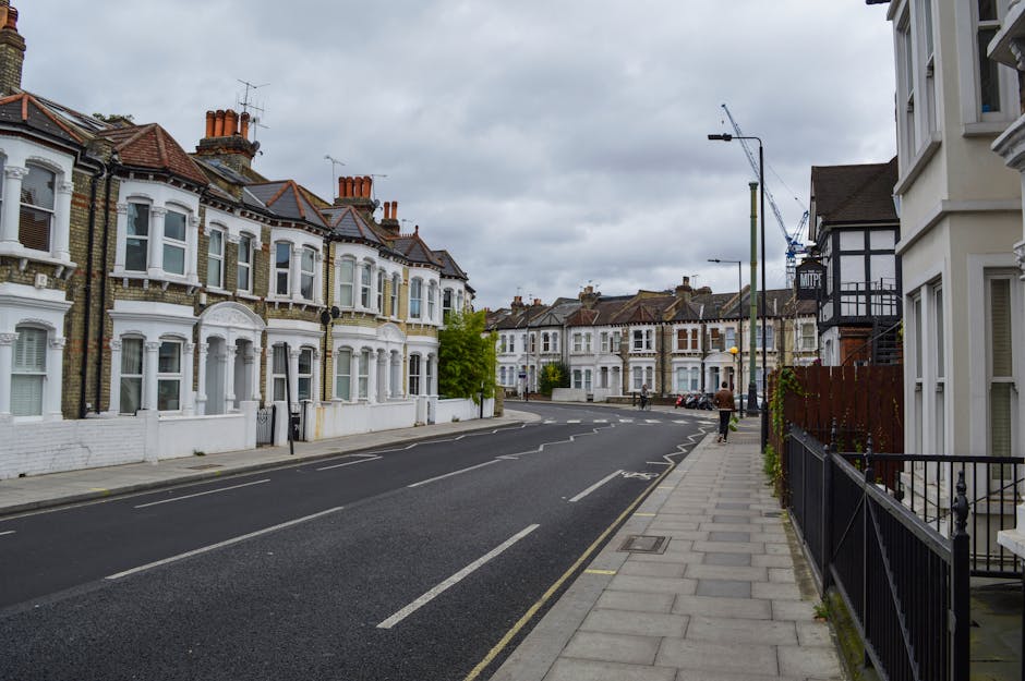 A wide view of a residential street in Harrow with Victorian-style terraced houses featuring bay windows and brick facades. The houses are painted in light colors, predominantly white, and have tiled roofs with multiple chimneys. The street is lined with a sidewalk paved with concrete slabs, bordered by black metal railings on the right side. There is a person walking along the sidewalk, and several parked cars are visible in the distance. Overhead, a large crane is visible, indicating construction activity nearby. The sky is overcast with grey clouds, creating a subdued lighting environment. This scene captures the typical urban setting where [COMPANY_NAME] provides house removals and furniture transport services, emphasizing moving logistics within Harrow’s residential areas.