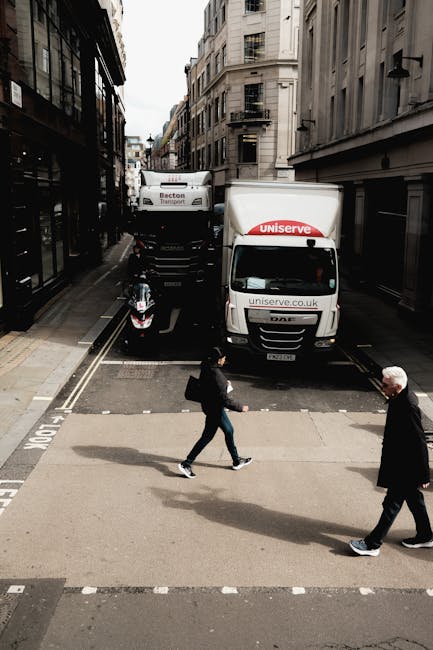 A narrow urban street scene during daytime showing a moving company's van parked in the middle of the road, with the logo 'uniserve.co.uk' and the word 'Uniserve' on its side. Behind the van, a larger truck with the label 'Becton Transport' is also parked, partially blocking the street. On the sidewalk, two pedestrians are crossing; one is a woman with a backpack and casual clothing, and the other is an older man wearing a dark coat and white hair. The street is flanked by multi-storey buildings with multiple windows and fire escapes, typical of city architecture. The road markings and signage indicate a designated loading or parking area for moving vehicles, supporting house removals and furniture transport activities. Natural daylight illuminates the scene, highlighting the urban environment and the busy activity typical of home relocation logistics.