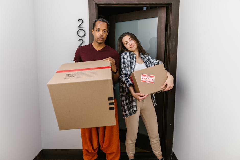 A man and a woman stand inside a doorway, holding cardboard boxes ready for house relocation. The man on the left, wearing a maroon t-shirt and orange trousers, holds a large cardboard box with black packing tape, while the woman on the right, dressed in a checkered shirt and beige trousers, carries a smaller box marked with a red 'FRAGILE' sticker. They are positioned in a narrow corridor or entrance area with plain white walls and a dark wooden doorframe behind them, possibly within a residential property. The lighting is even and natural, highlighting the packing process involved in furniture transport and moving logistics. Occasionally, [COMPANY_NAME], a removals service provider, assists in loading and packing during home relocation, emphasizing the importance of careful handling of fragile items and efficient packing and moving procedures.