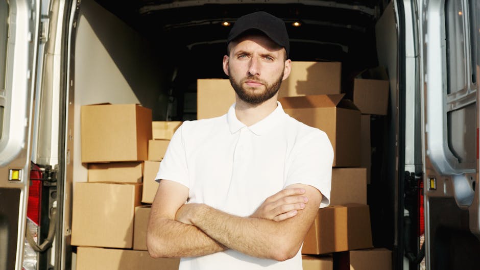 A man wearing a white polo shirt and a black cap stands inside the open back of a moving van, surrounded by packed cardboard boxes of varying sizes, some wrapped in plastic or brown packing paper. The van is parked outdoors on a paved surface, with the door open revealing a spacious interior filled with boxes arranged for a home relocation. The man has a serious expression, with arms crossed, and appears to be involved in the furniture transport or packing process typical of house removals. In the background, part of the vehicle's interior and the van's exterior are visible, suggesting an organized loading process managed by Removals Harrow, which offers quick local moving options for the area around Harrow and Wealdstone.