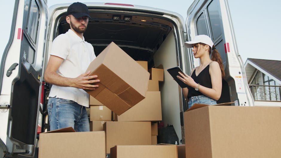 A man and a woman are positioned near the open back of a white moving van, which is parked outdoors in a residential area, with a house visible in the background. The man, wearing a black cap, white polo shirt, and jeans, is holding a medium-sized cardboard box as part of a home relocation process. The woman, wearing a white cap, black tank top, and jeans, is standing nearby, focused on a tablet device in her hands. Inside the van, several packed cardboard boxes of varying sizes are stacked, some on the floor and others against the interior walls. The vehicle's interior appears spacious, suitable for furniture transport, and the scene captures an active loading process involving careful handling of packing materials. The lighting is natural, indicating daytime, and the environment suggests an organized move managed by Removals Harrow, specializing in local house removals and furniture transport, with attention to packing and moving logistics to ensure a smooth relocation.