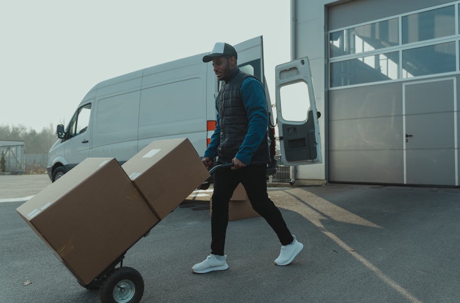 A man wearing a white polo shirt and a black cap stands inside the open back of a moving van, surrounded by packed cardboard boxes of varying sizes, some wrapped in plastic or brown packing paper. The van is parked outdoors on a paved surface, with the door open revealing a spacious interior filled with boxes arranged for a home relocation. The man has a serious expression, with arms crossed, and appears to be involved in the furniture transport or packing process typical of house removals. In the background, part of the vehicle's interior and the van's exterior are visible, suggesting an organized loading process managed by Removals Harrow, which offers quick local moving options for the area around Harrow and Wealdstone.